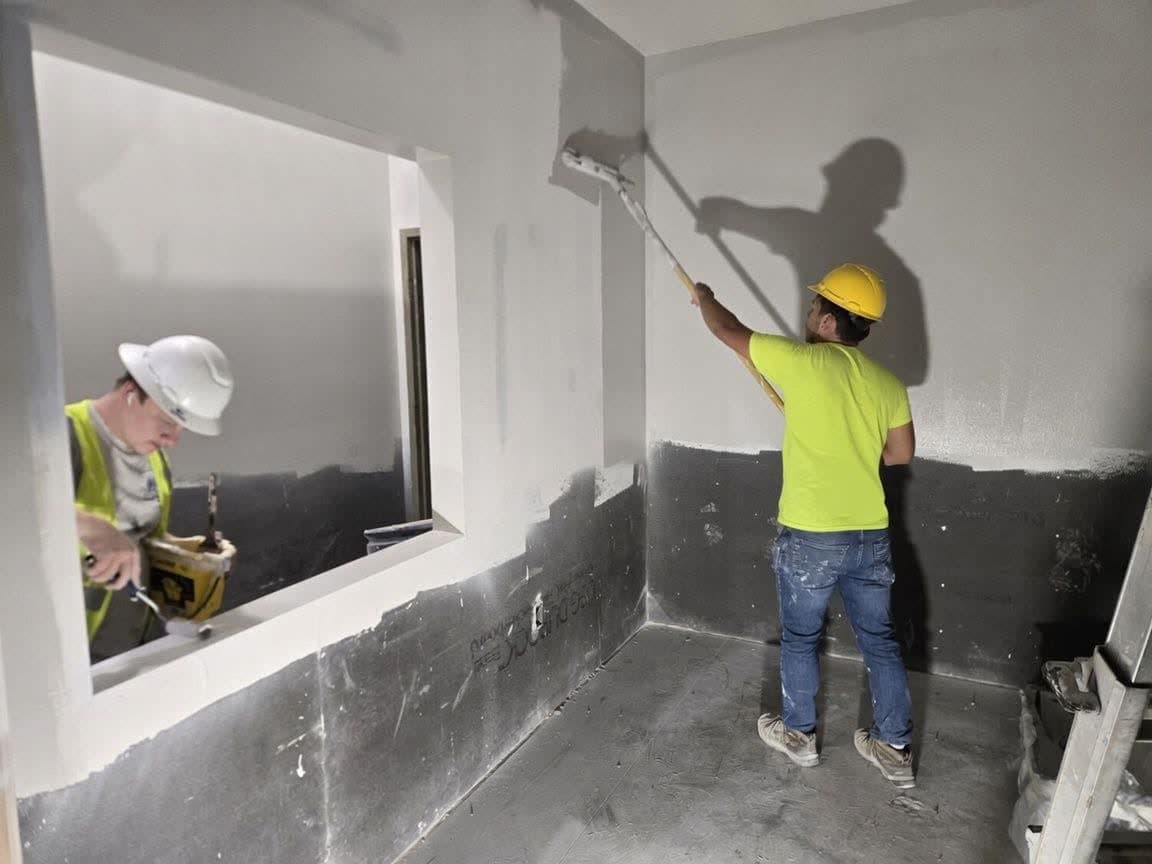 Two workers in hi-vis and hard hats, one rolling a wall with an extension pole on a commercial job